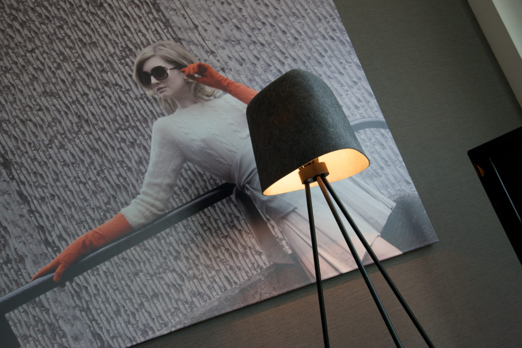 Grey Tom Dixon lamp on tripod in front of large photograph of woman in sunglasses, encouraging people to support green travel and turn off lights when they're not using them.